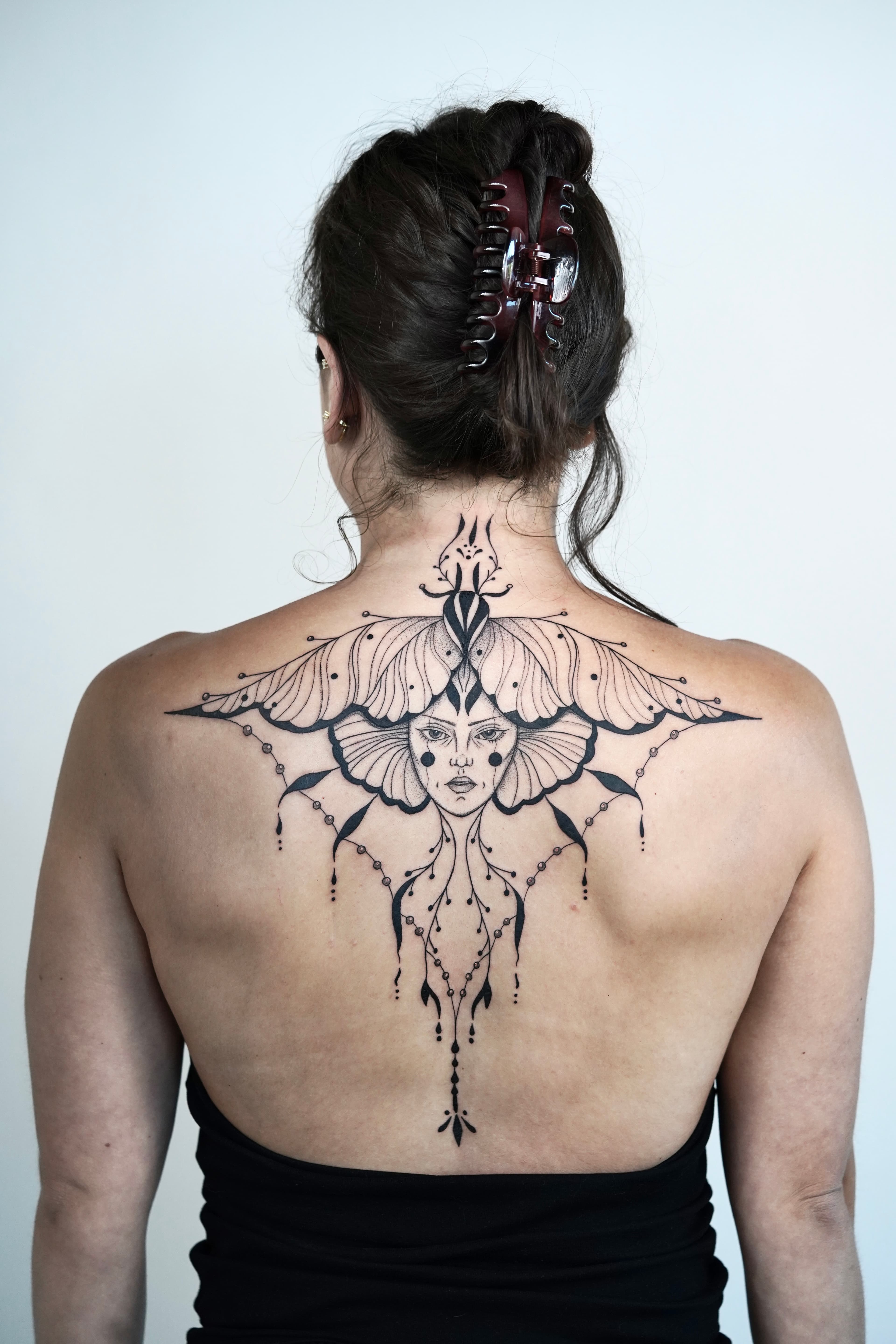 Fine line moth and feminine portrait tattoo across the upper back by Nika Osseili.